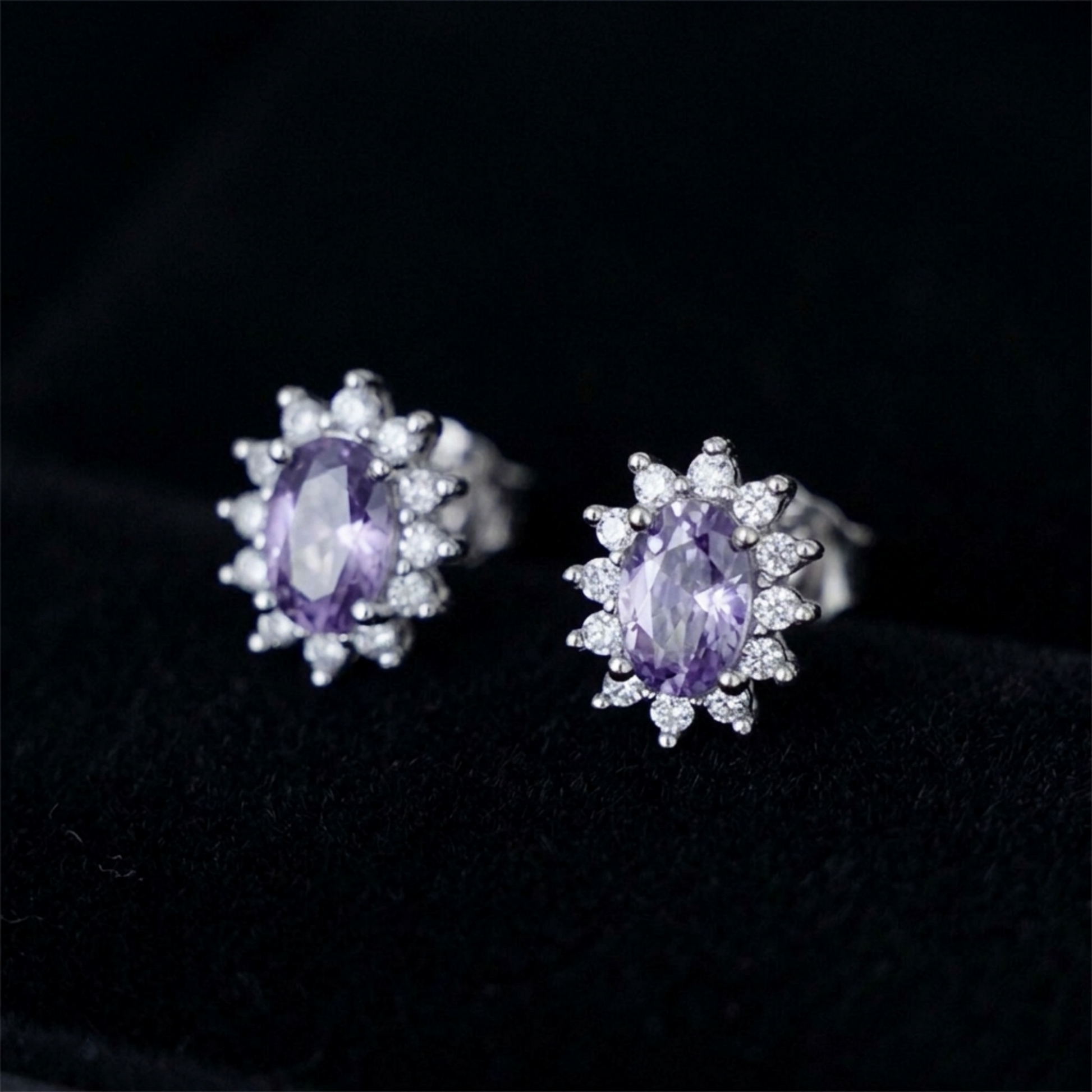 Close-up of oval amethyst cluster stud earrings with white stone halo, silver-toned setting