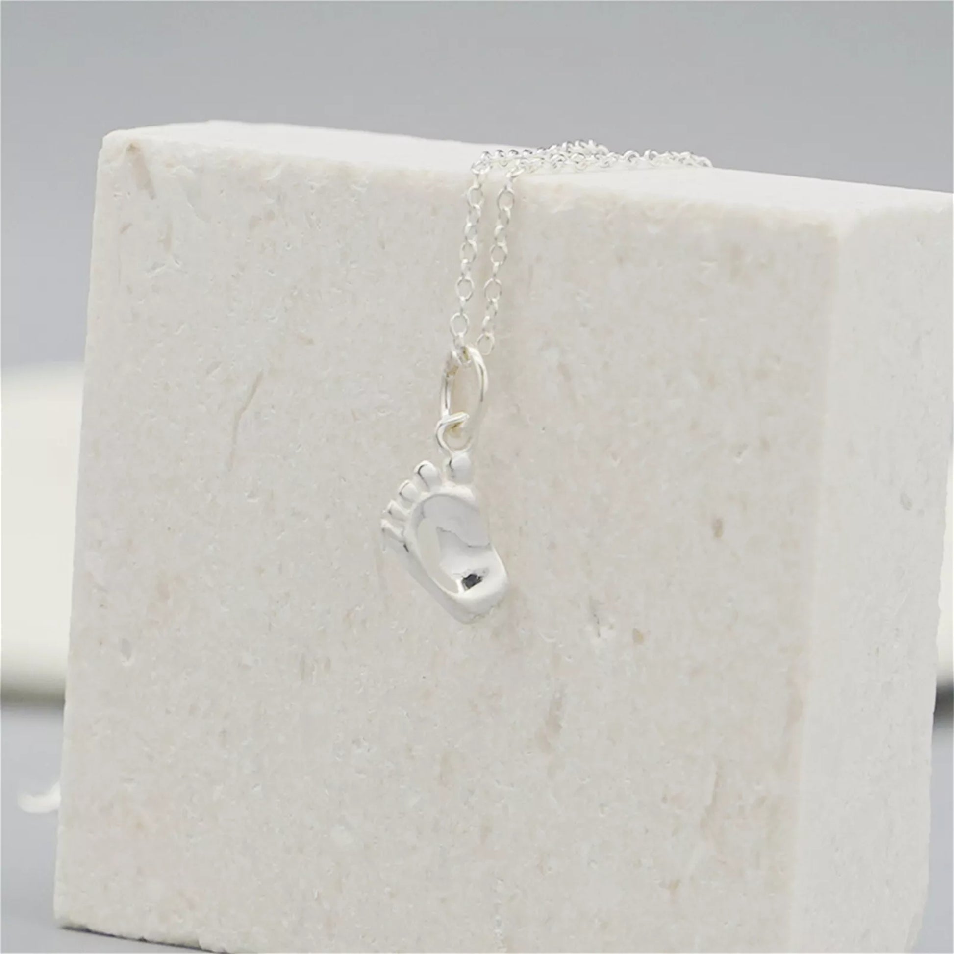 Silver baby foot charm pendant hanging from a chain on a white display block