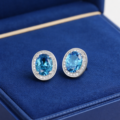 Blue topaz CZ halo studs, angled view: oval blue stone with clear halo edge, silver posts.