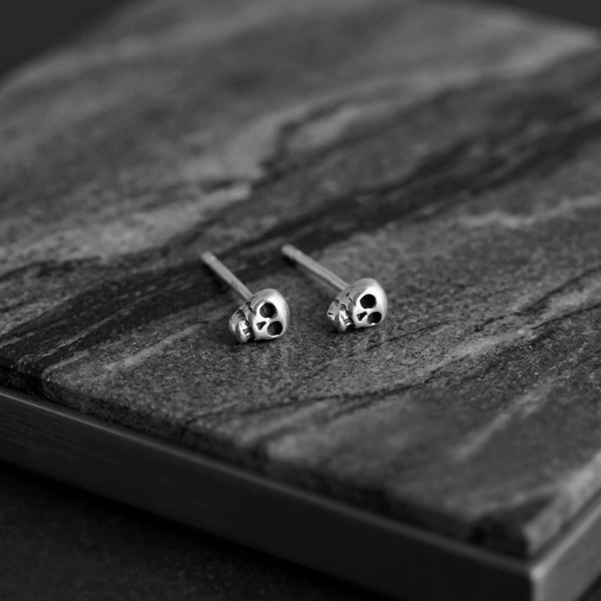 Skull stud earrings resting on a surface; small black-eyed skull details, silver-tone