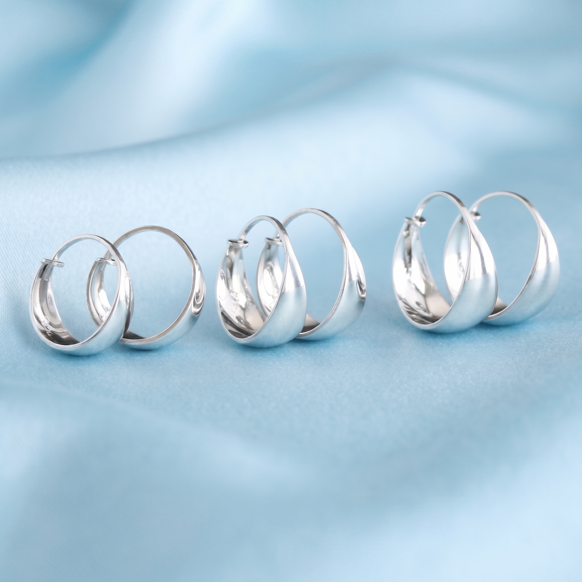 Silver wide dome huggie hoop earrings shown from different angles on soft blue fabric