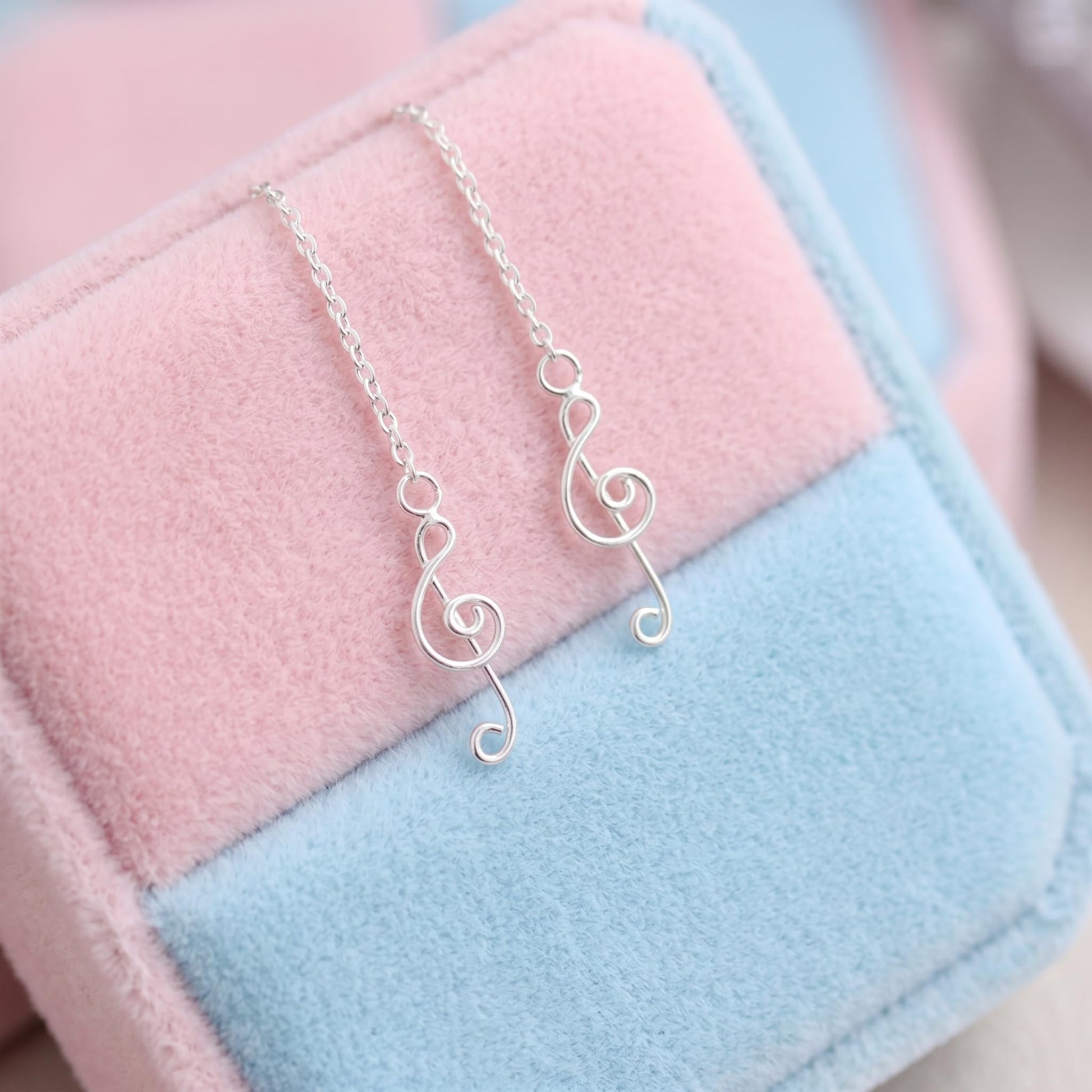 Silver treble-note threader dangle earrings resting on pastel pink and blue fabric